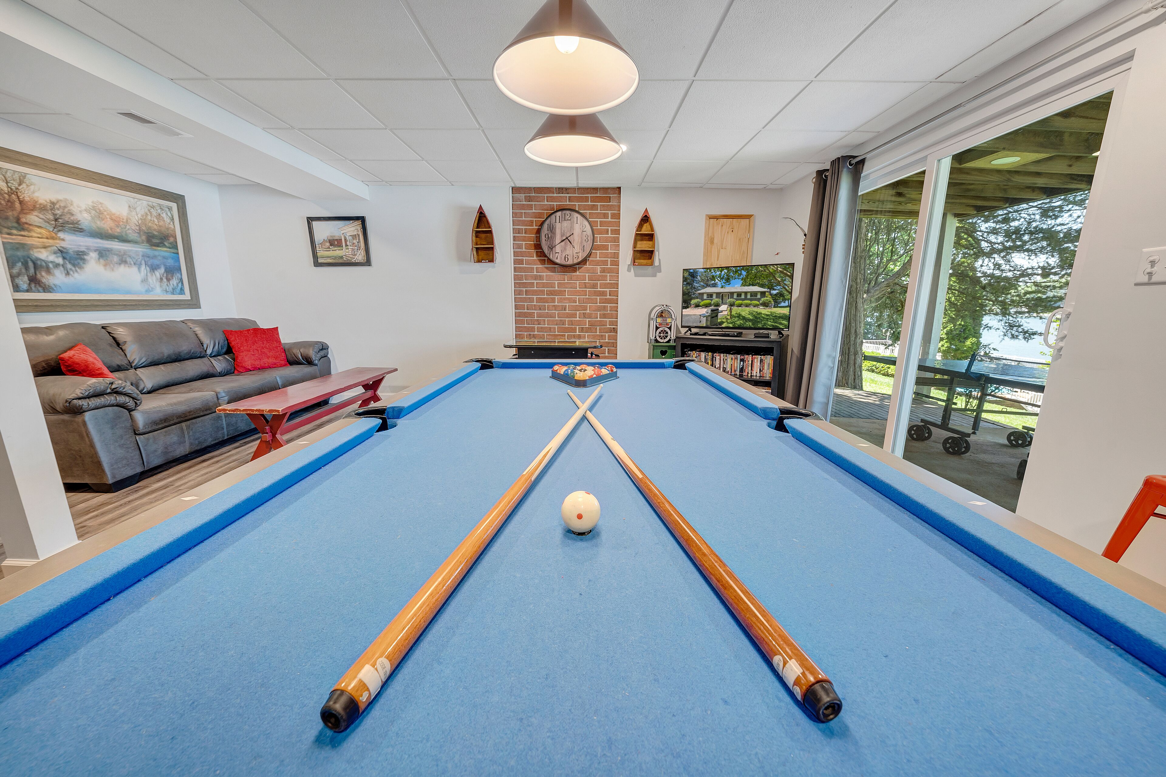 A pool table with two cues and a ball, a sofa with red cushions, a clock on the wall, and a TV in the background.