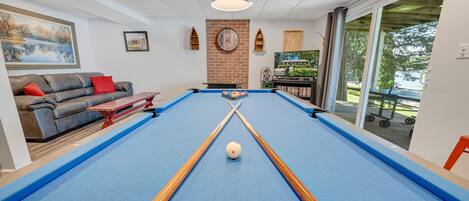 A pool table with two cues and a ball, a sofa with red cushions, a clock on the wall, and a TV in the background.
