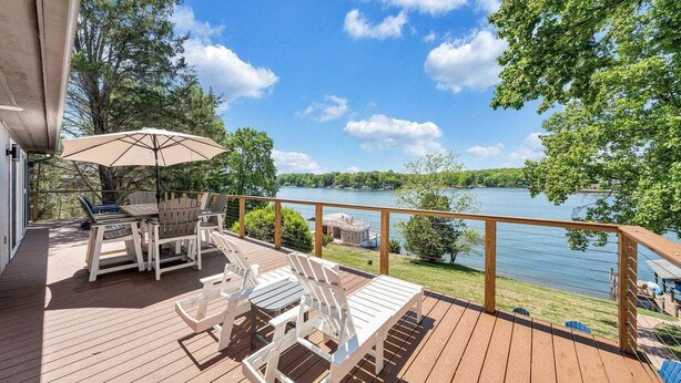 A wooden deck with outdoor furniture, a table, and chairs, overlooking a lake with trees and a clear sky. - Lakeside Sunsets - Private Dock, Hot Tub & Firepit (Moneta)