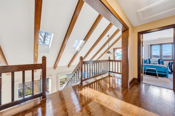 Una habitación amplia con techos altos y vigas de madera a la vista, una escalera que lleva a un nivel superior y vistas al exterior a través de amplios ventanales.