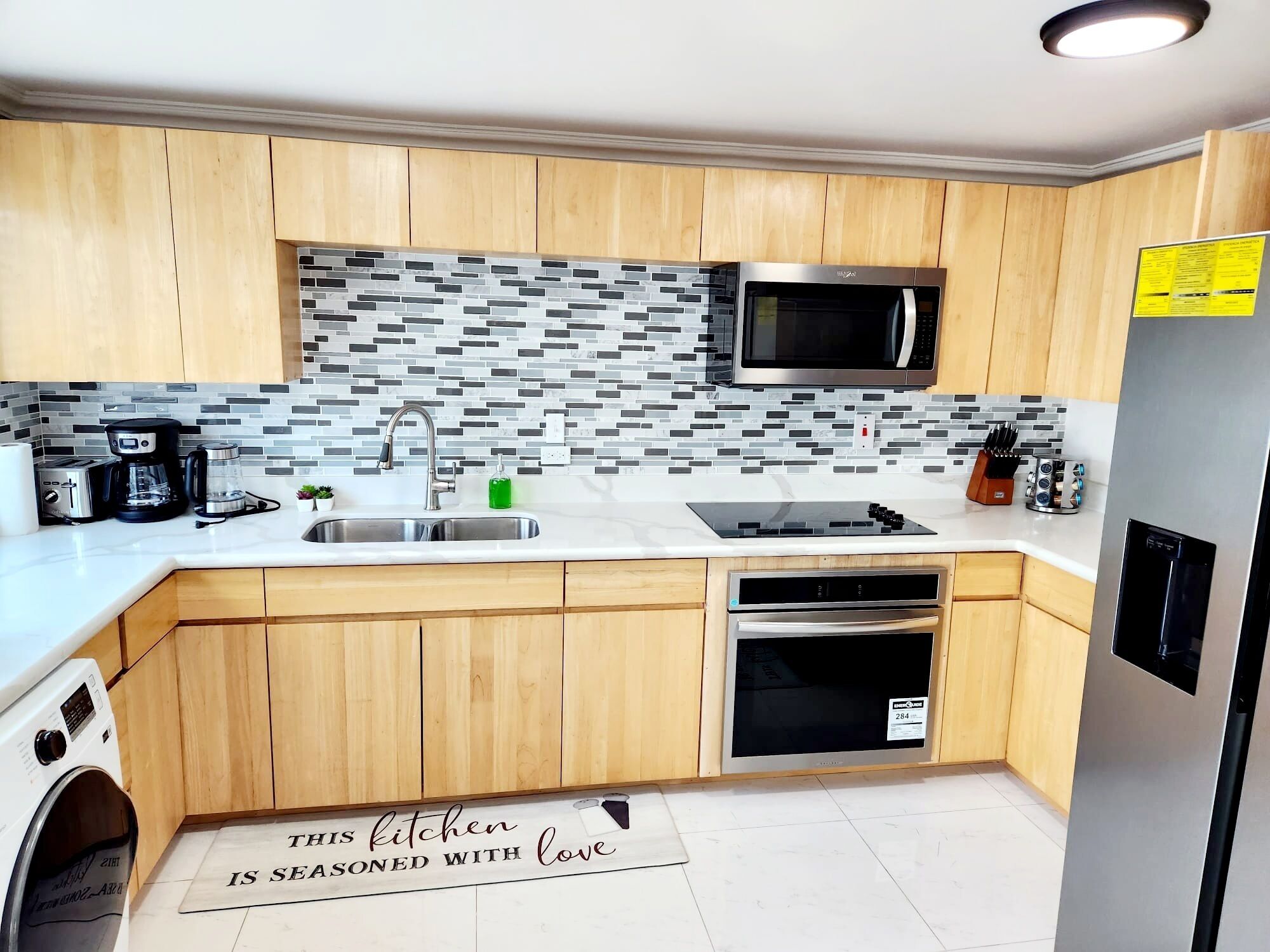 Une cuisine moderne avec des meubles en bois, un pare-feu en carrelage, des appareils en acier inoxydable et un tapis de cuisine orné d’un message.