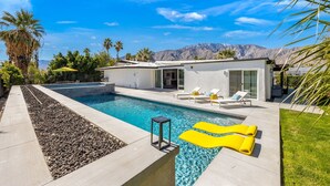 Ein moderner Poolbereich mit freiem Blick auf Berge im Hintergrund, eine Terrasse mit Liegestühlen und ein kleines Gebäude mit großen Fenstern.