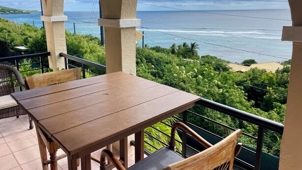 Un balcon avec une table et des chaises en bois, offrant une vue sur la côte, l’océan et la végétation.