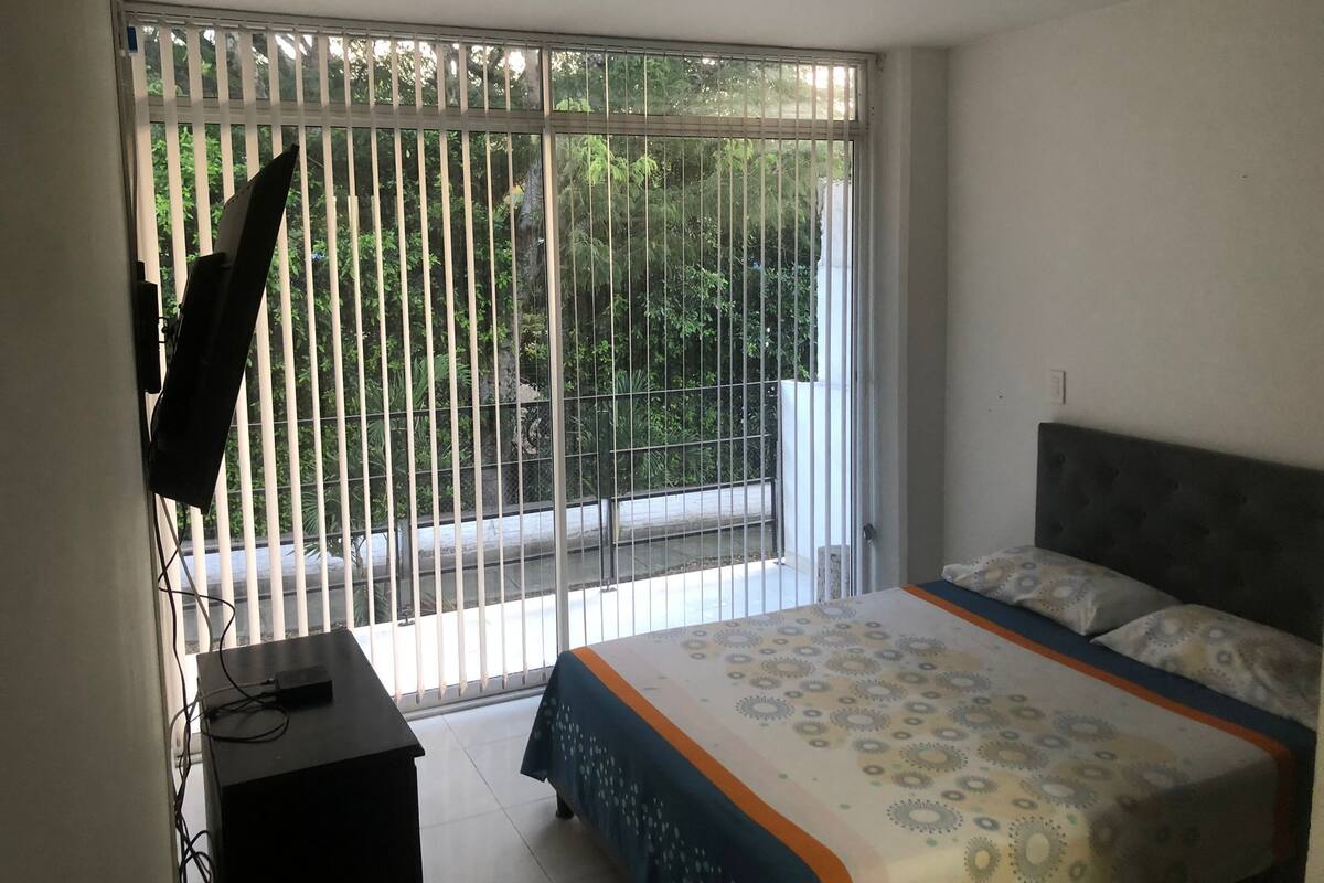 Een slaapkamer met een bed, een aan de muur bevestigde televisie en een balkon met uitzicht op groen.