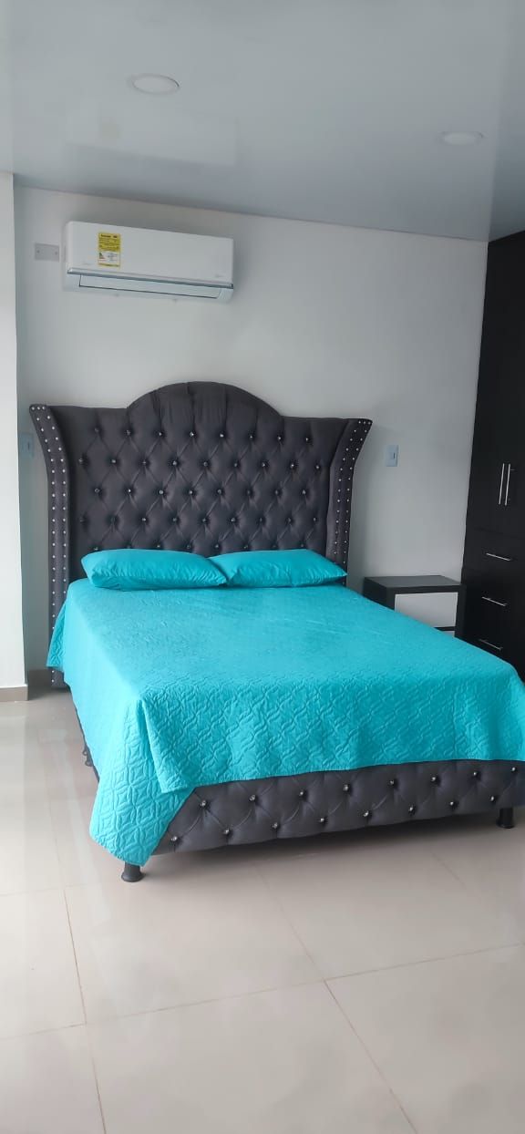 Una cama bien hecha con un cabecero oscuro y ropa de cama color verde azulado en una habitación con piso de baldosas y un armario oscuro.