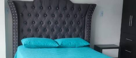 Una cama bien hecha con un cabecero oscuro y ropa de cama color verde azulado en una habitación con piso de baldosas y un armario oscuro.