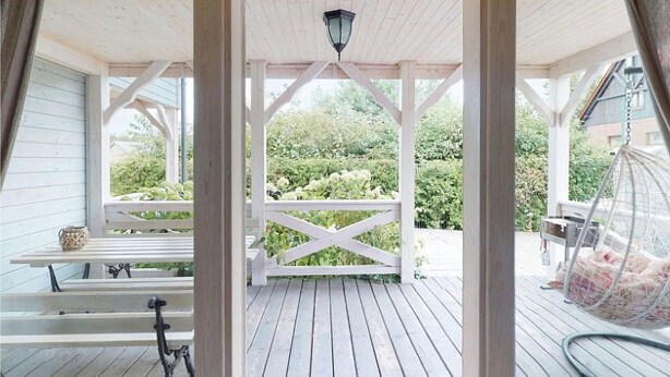 En overdækket veranda med en træbænk, en hængende fuglefoder og udsigt til grøn vegetation.