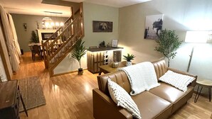 Uma sala de estar com sofá de couro marrom, piso de madeira, escada, espaço de refeições e cozinha.