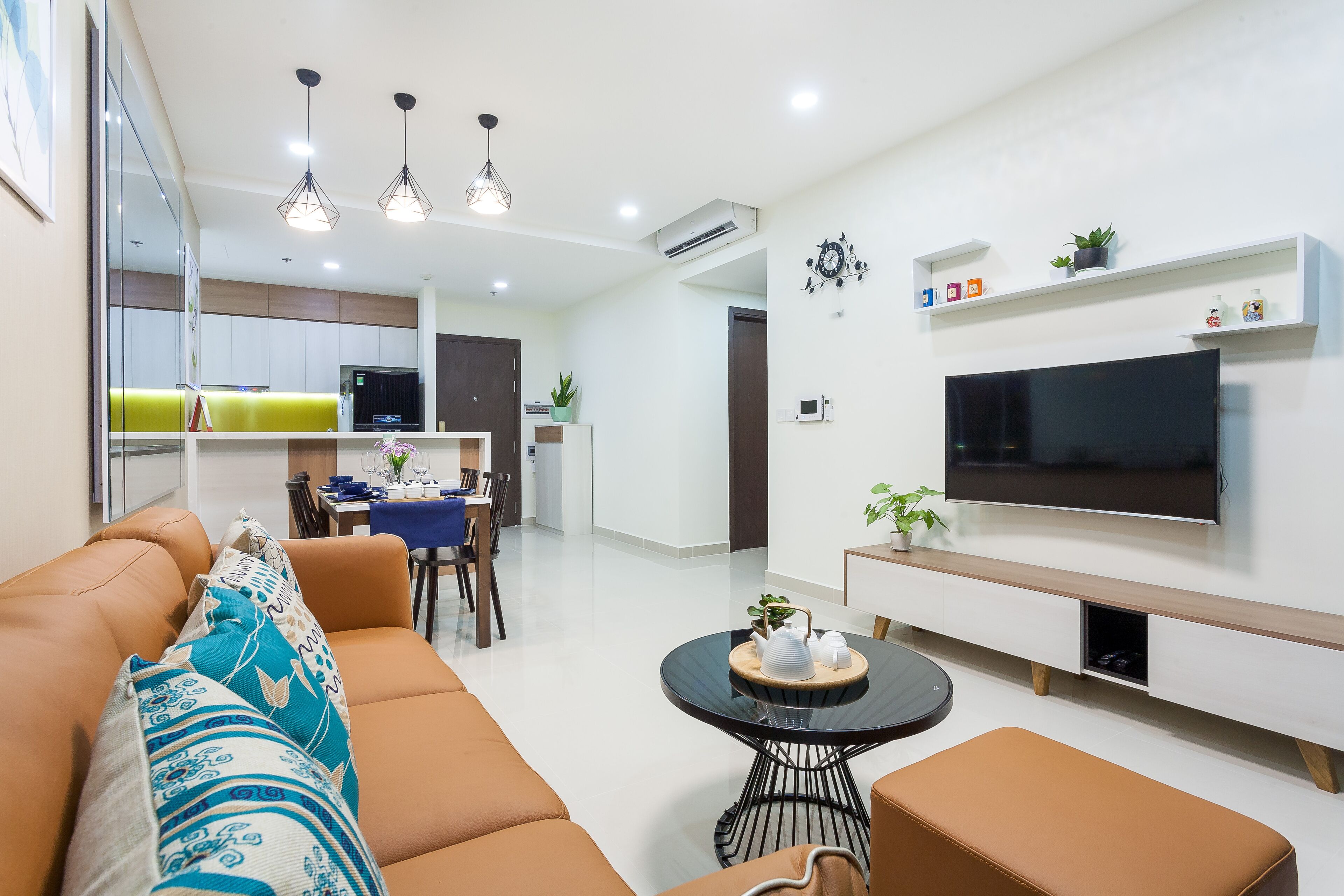 A modern living room with a sofa, a coffee table, and a TV mounted on the wall. There is a dining area in the background with a table and chairs.