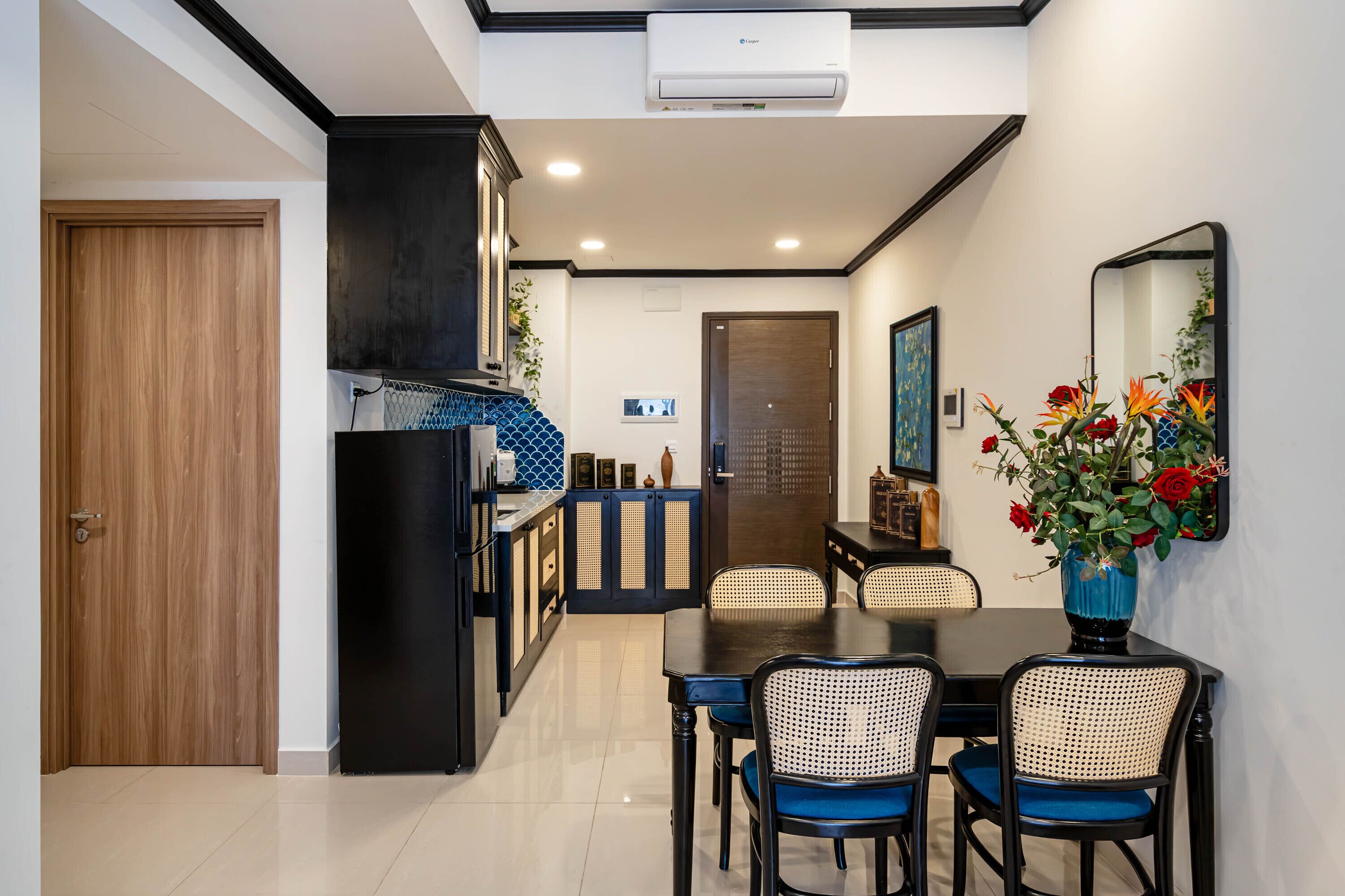 Una cocina moderna con armarios oscuros, un comedor con una mesa negra y sillas azules, y un jarrón decorativo con flores en la pared.