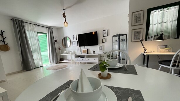 A modern dining area with a white table set for two, a television mounted on the wall, and a desk with a lamp. - Buganvilla house, super wifi, sunny terrace (Playa Blanca)