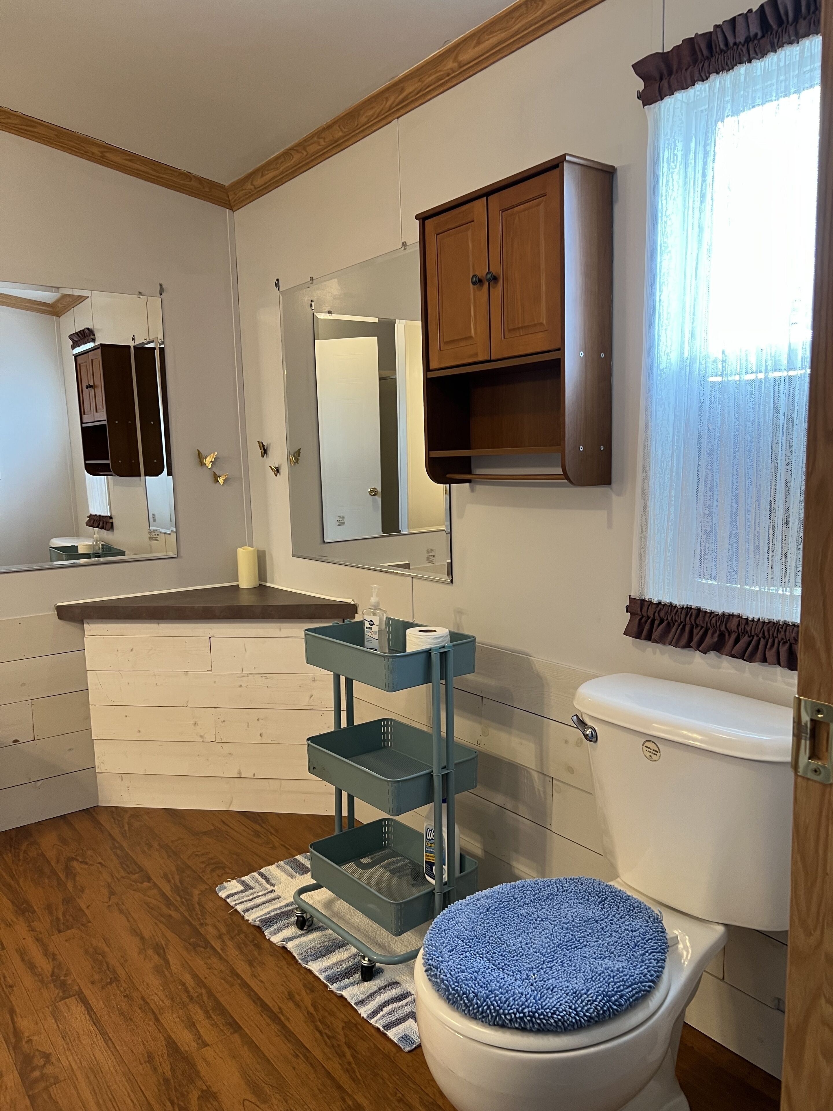 A bathroom with a white toilet, a blue toilet seat, a mirror, a wooden cabinet, a sink with a mirror, and a window with curtains.