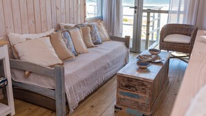 Ein hölzernes Bett mit weißer Bettdecke und mehreren Kissen, ein Korbstuhl mit Sitzkissen sowie ein kleiner Holztisch mit Schalen.