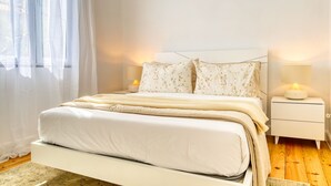 Un dormitorio con cama, cortinas blancas, una lámpara de noche y una mesita de noche.