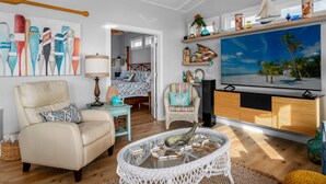 A living room with a large TV, a white sofa, a wicker coffee table, and a colorful painting on the wall.