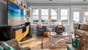 A living room with a large TV, wooden cabinets, blue armchairs, a round coffee table, and a view of the ocean through large windows.