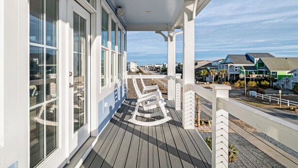 A covered porch with rocking chairs, large windows, and a view of the beach and houses.