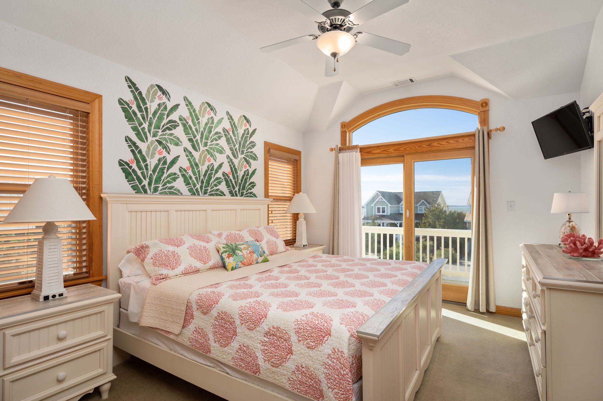 A bedroom with a bed, two bedside tables, a ceiling fan, a large window with blinds, a view of the outside, and a mounted TV.