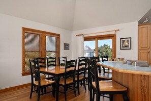A dining area with a wooden table and chairs, large windows with blinds, and a view of a house outside.