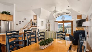 A dining area with a wooden table, chairs, and a plant centerpiece. A staircase with wooden railings leads to a lofted living area.