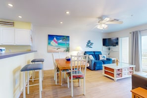 A modern living room with a dining area, a blue sofa, a TV mounted on the wall, and a ceiling fan.