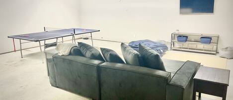 A room with a large grey sofa, a ping pong table, and a small wooden table.