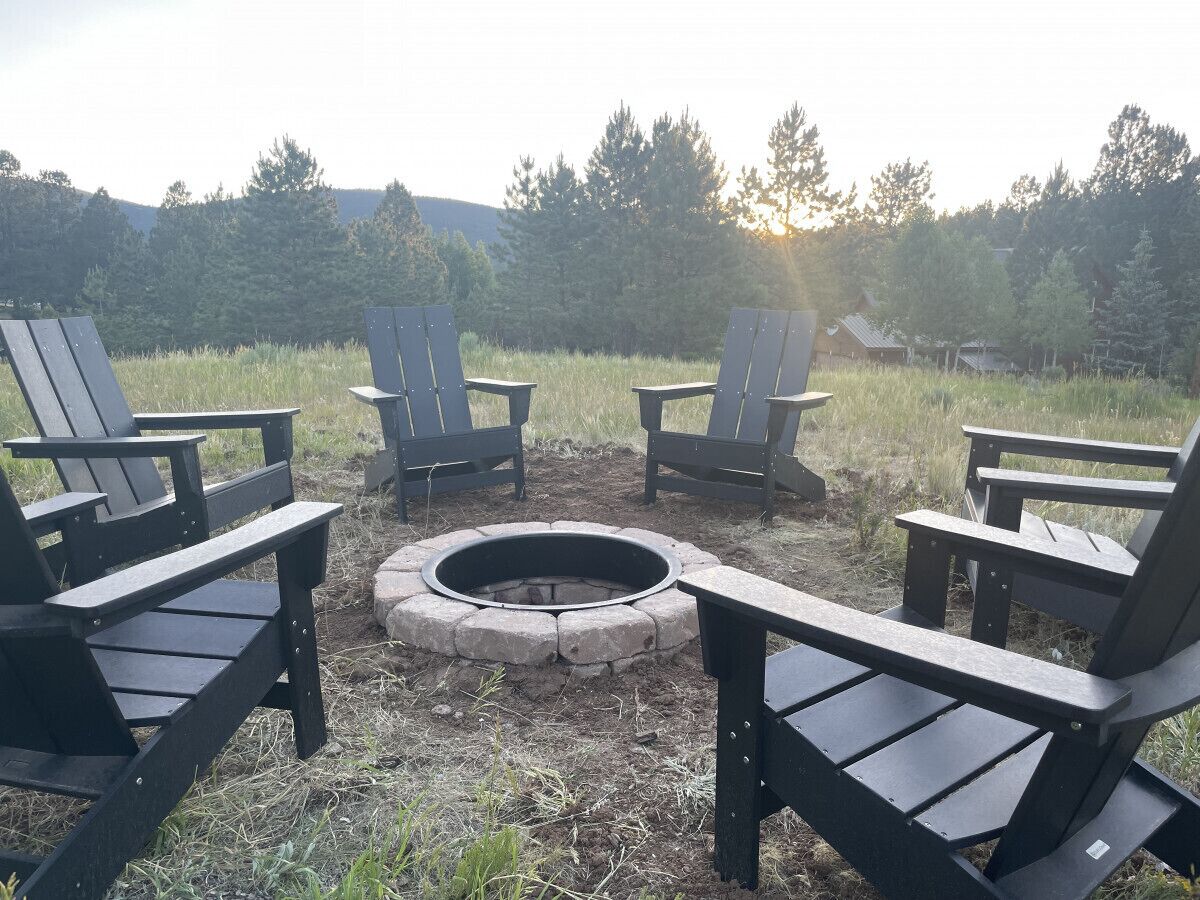 Uma fogueira circular rodeada por quatro cadeiras Adirondack em ambiente externo, com um campo gramado e árvores ao fundo.