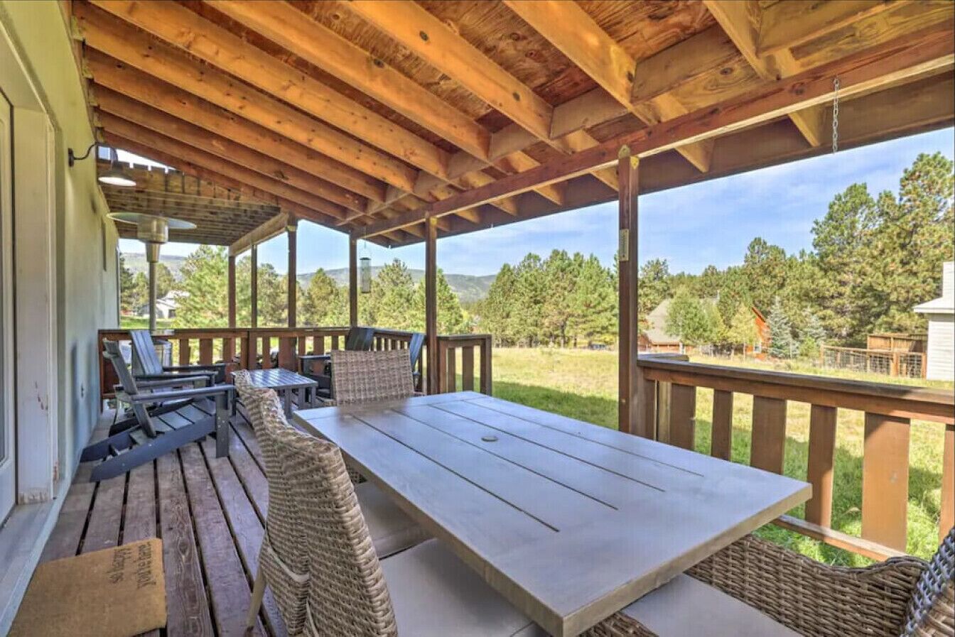 A covered outdoor patio with a wooden table and wicker chairs, overlooking a grassy area with trees.