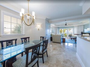 A spacious dining area with a glass table and dark wooden chairs, a chandelier, and a view of the living room.