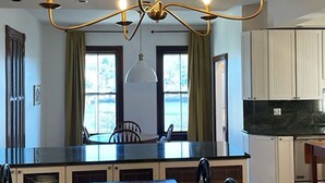 A dining area with a long wooden table, six chairs, a chandelier, and a view of the kitchen through large windows. - Amazing experience near the Quinnipiac River. Historic w/chef's kitchen & bidet. (New Haven)