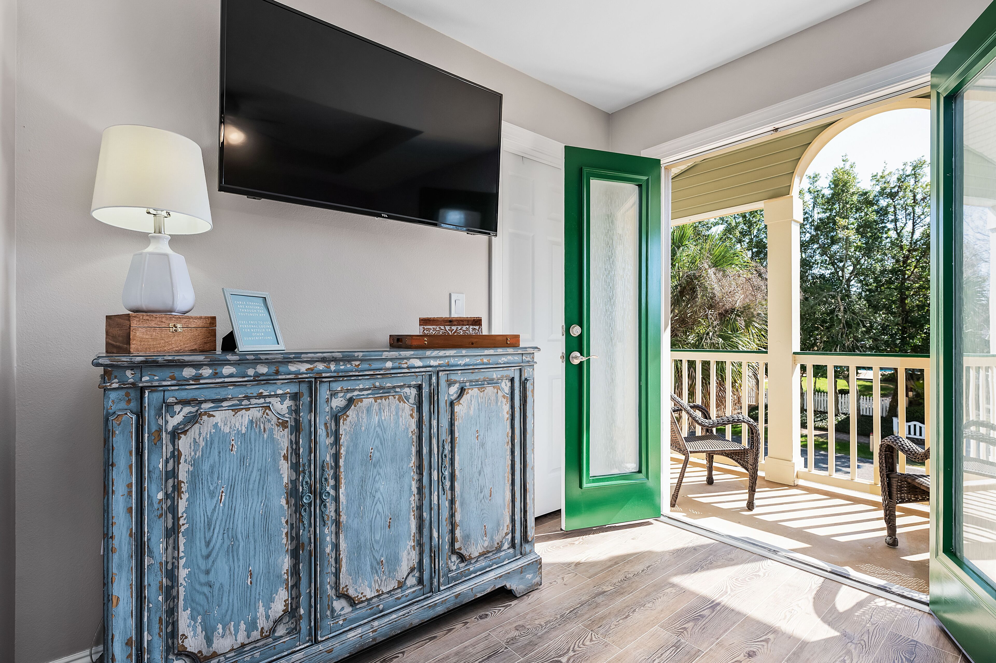 Una habitación con un armario de madera, un televisor montado y una puerta verde que da a una terraza exterior con sillas.