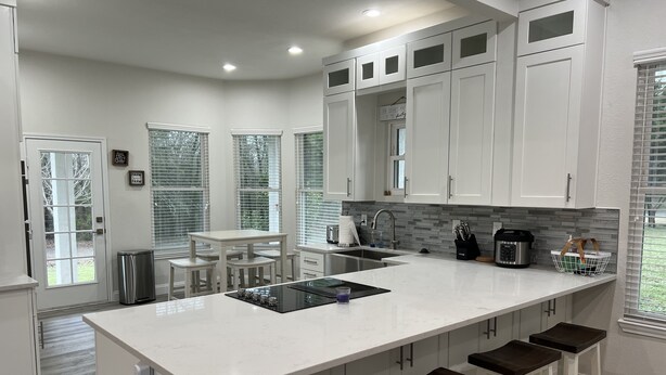 A modern kitchen with white cabinets, a central island with stools, a built-in stove, and a large window with blinds.