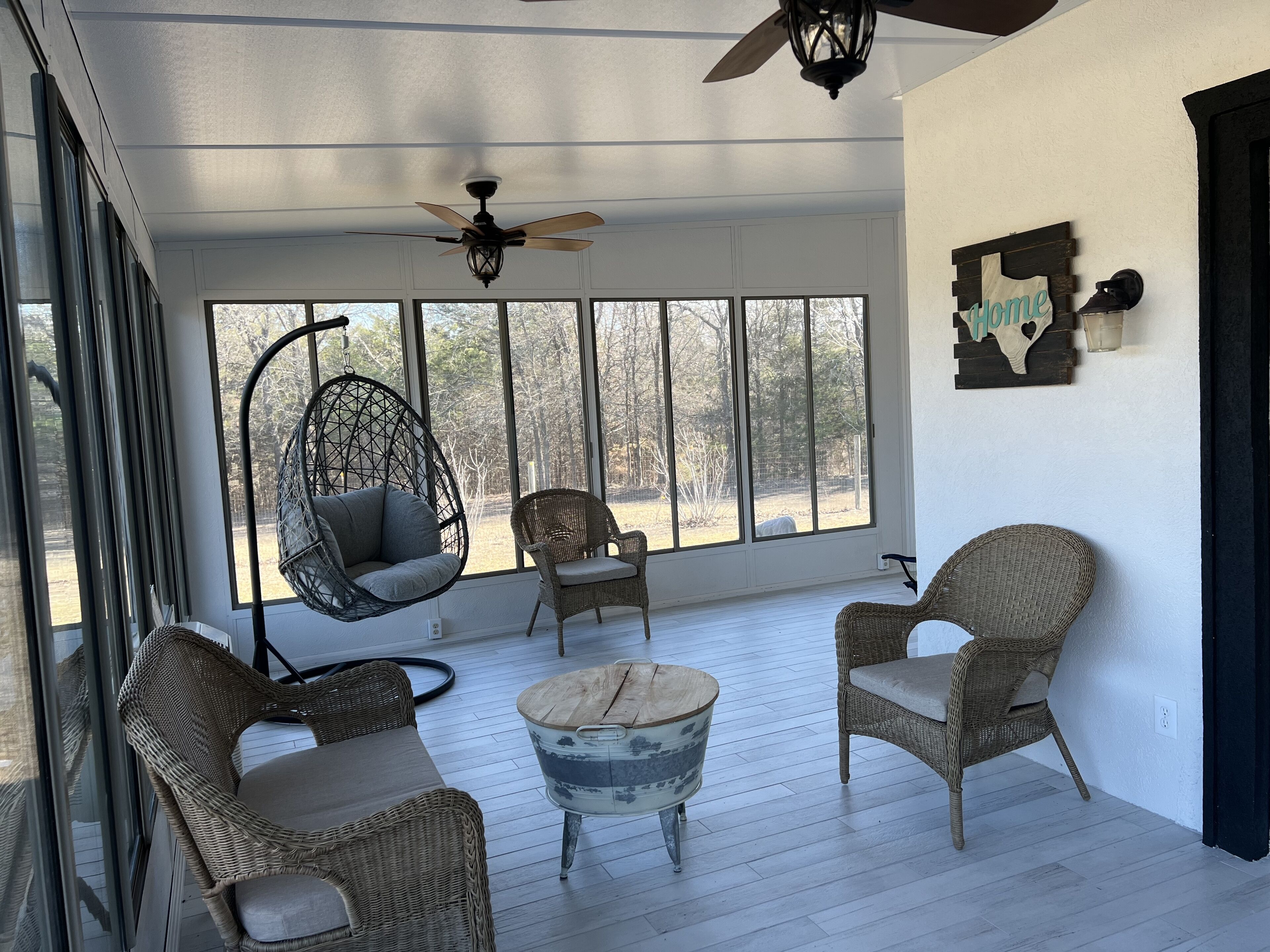 A sunlit room with wicker furniture, a ceiling fan, and large windows offering a view of the outdoors.