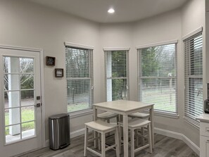 A corner kitchen with a white table and two barstools, large windows with blinds, a trash can, and a door leading outside. - Experience Elegance at our Luxury Ranch with New Pool & Deck (Celina)