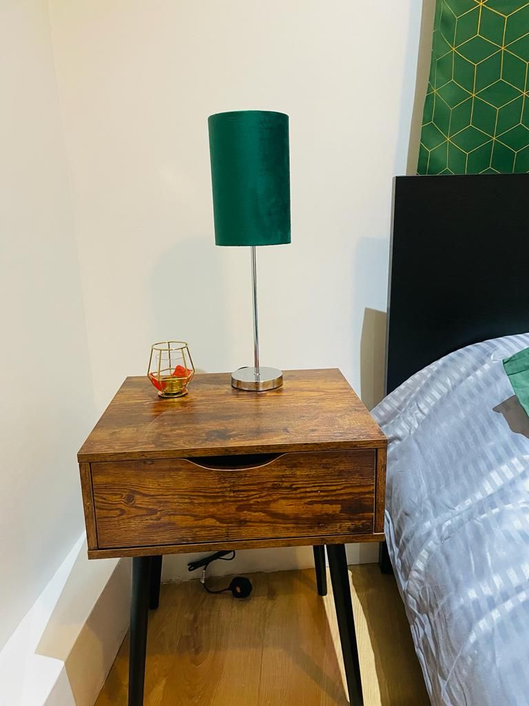 Ein Nachttisch aus Holz mit einer grünen Lampe, einer Glasbowl und einer Schublade.