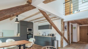 En rummelig loftsuite med synlige træbjælker, et tekøkken, et spisebord med stole og en trappe til en etage over.