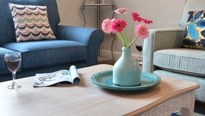 Un salón con un sofá azul, una mesita con un jarrón de flores rosas, una copa de vino tinto y una revista.