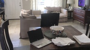 A living room with a dining table, a laptop, papers, and a bowl. There is a sofa, a chair, and a TV in the background.