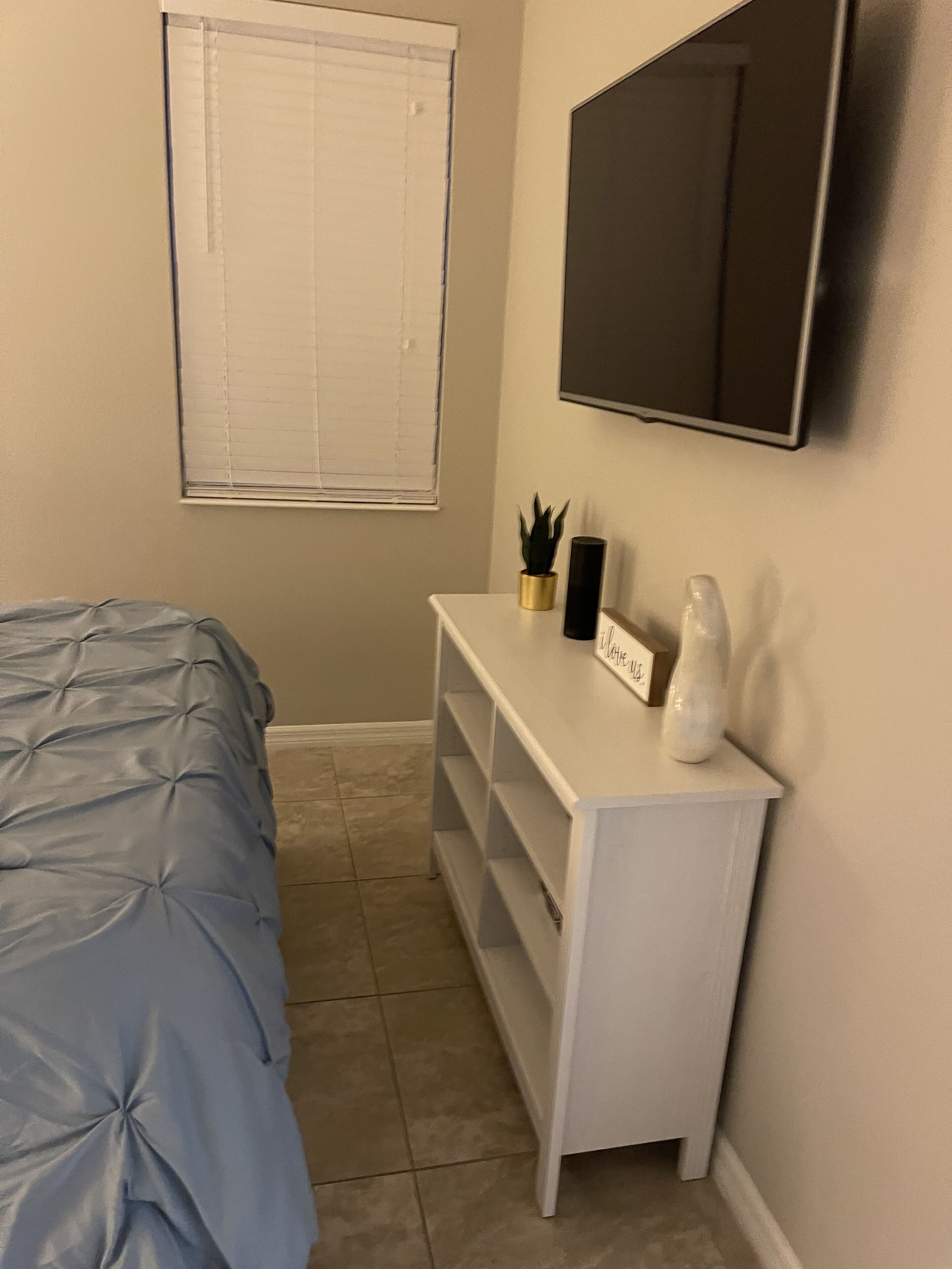 Ein Schlafzimmer mit einem Bett, einem an der Wand montierten Fernseher, einem kleinen weißen Schminktisch mit Pflanze und einem Fenster mit Jalousien.