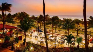 A beachfront restaurant with outdoor seating, palm trees, and a view of the ocean at sunset.