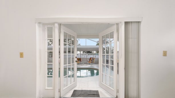 Une vue à travers une porte vitrée donnant sur un espace piscine, avec un patio couvert et du mobilier d’extérieur.