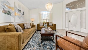 Un salon comprenant deux canapés, une table basse et un lustre. Il y a une grande fenêtre avec des rideaux et une porte ornée d’un panneau de verre décoratif.