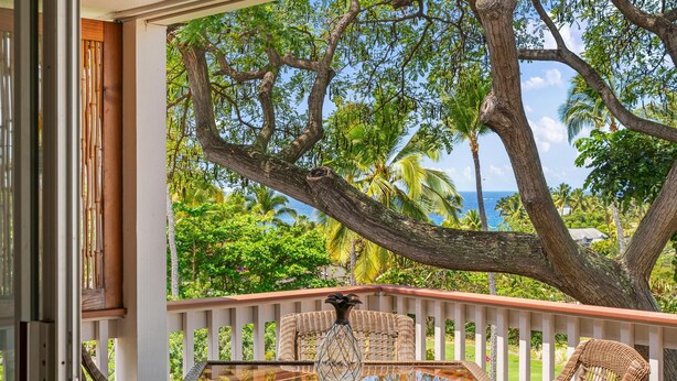 A glass-top table with wicker chairs and a decorative vase on a balcony with a white railing, overlooking a tropical landscape with palm trees and a view of the ocean. - Oceanview Townhome, freshly updated in Keauhou, close to Kona. (Keauhou)