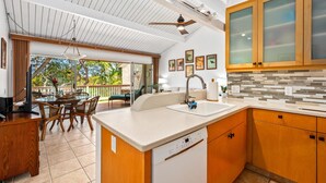 A modern kitchen with wooden cabinets, a dishwasher, and a dining area with wicker chairs and a glass table. - Oceanview Townhome, freshly updated in Keauhou, close to Kona. (Keauhou)