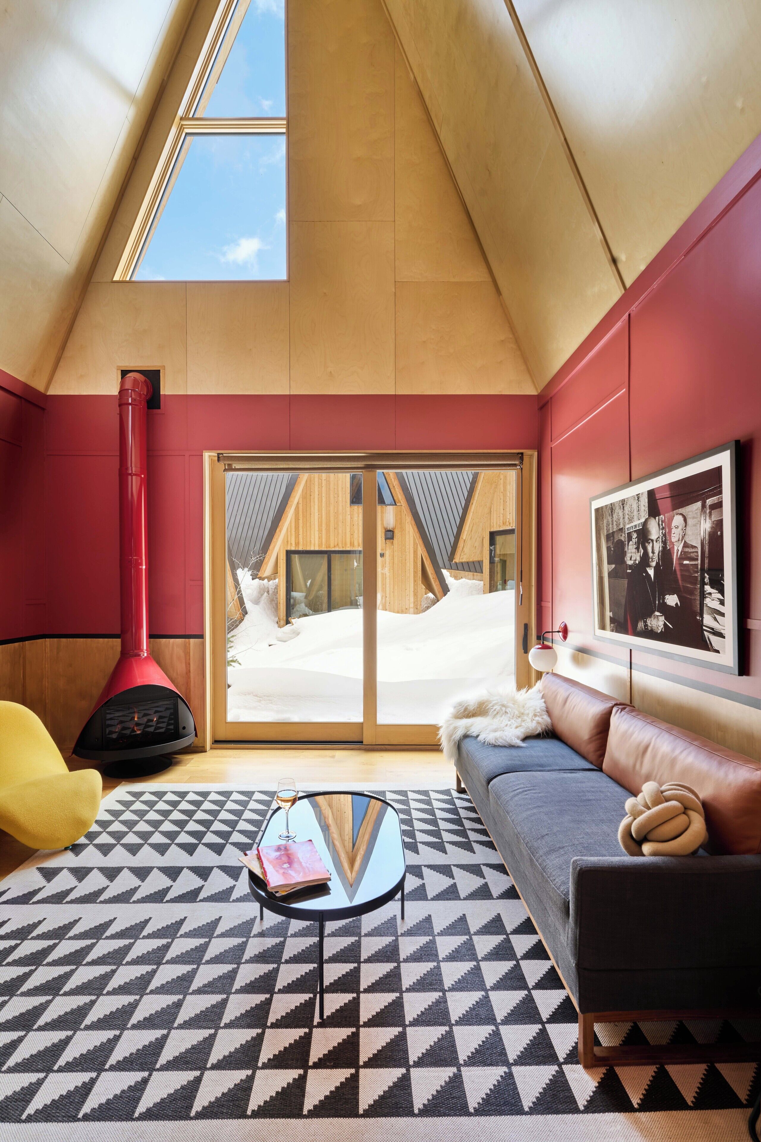 A cozy room with a sloped ceiling, a red wood-burning stove, a sofa, a coffee table, and a large window with a view of a wooden cabin.