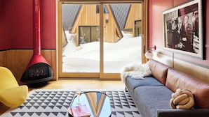 A cozy room with a sloped ceiling, a red wood-burning stove, a sofa, a coffee table, and a large window with a view of a wooden cabin. - A Frame Club  (Winter Park)