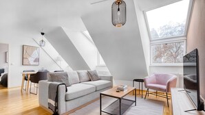 Un salon moderne avec un canapé gris, une table basse et un fauteuil rose. On y trouve un téléviseur posé sur un meuble et un luminaire suspendu au plafond.