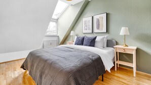 Une chambre avec un plafond en pente, un lit recouvert d’une couvre-lit à motifs, deux tables de chevet avec des lampes et deux œuvres d’art encadrées accrochées au mur.