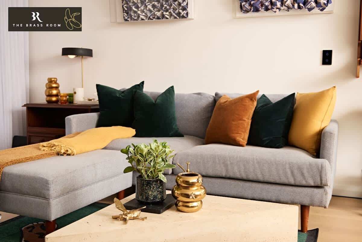 A modern living room with a grey sofa, multiple cushions, a small side table with a lamp, and decorative items.