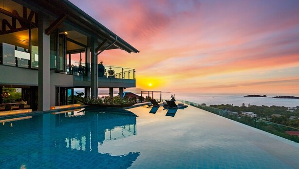 Una piscina infinita moderna con vistas al atardecer, una casa grande con mobiliario exterior y un cielo despejado.